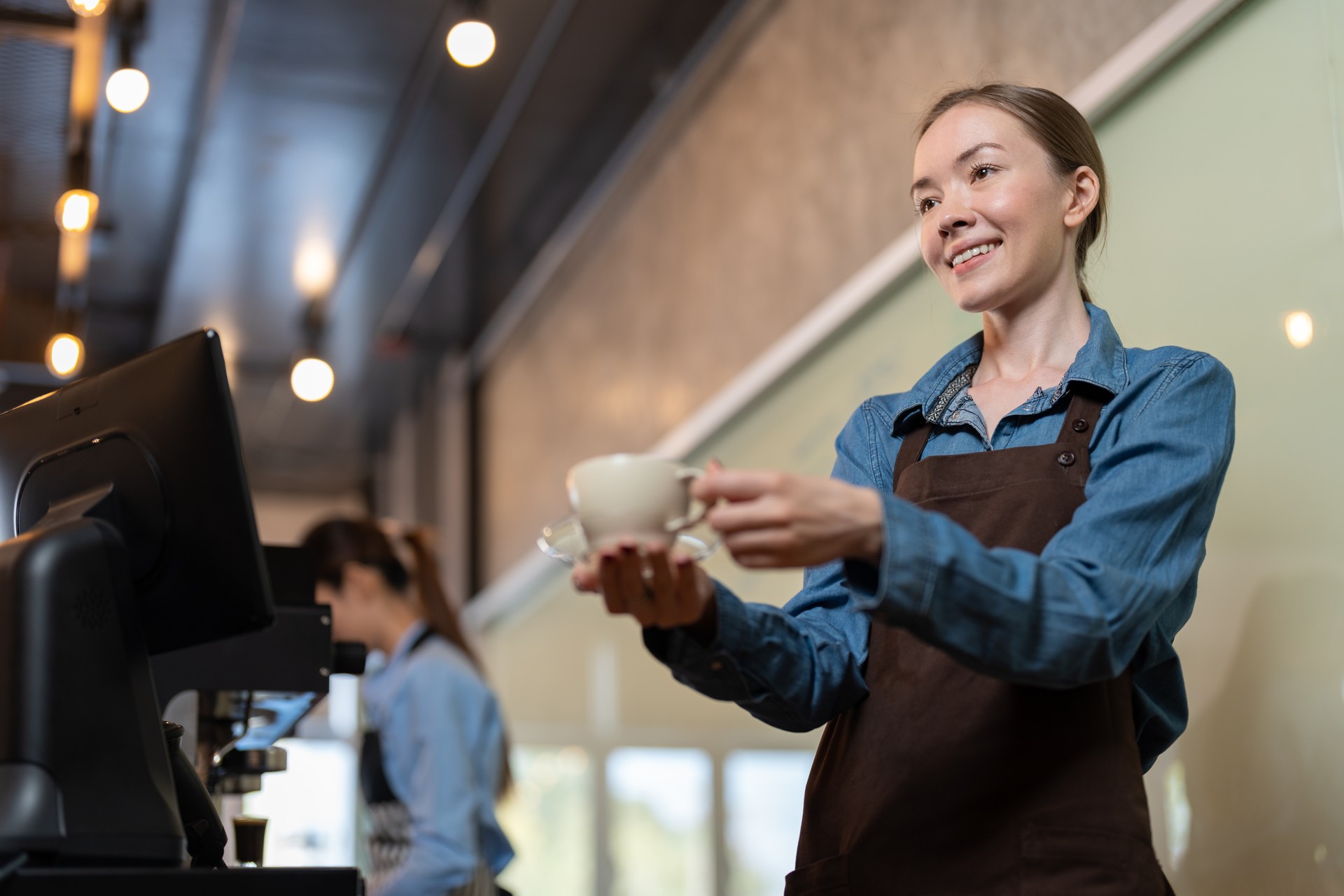 Female barista in apron smiling and serving cup of hot coffee to client in coffee shop. Barista owners small business.